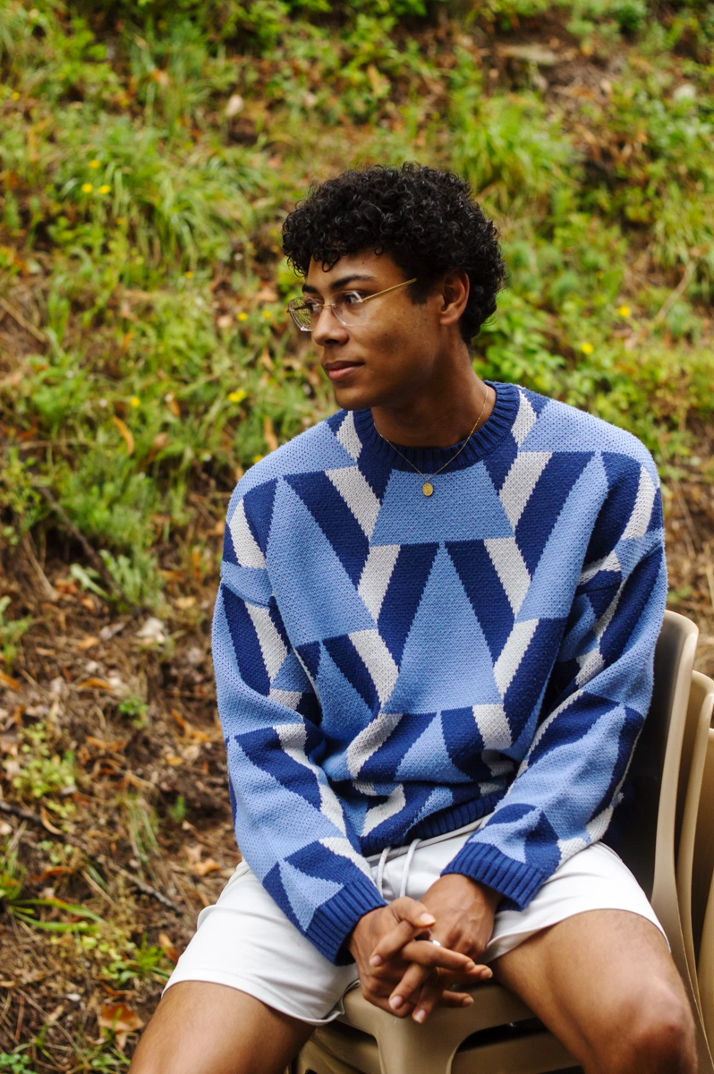 A young light-skin man from the side wearing a bright blue graphic pullover, sat on a chair, looking in the distance.